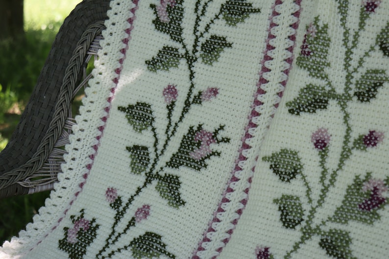 May include: A cream-colored, crocheted blanket with a floral cross-stitch design. The blanket features green leaves and pink flowers, with a scalloped edge and a border of pink triangles. The blanket is draped over a wicker chair.