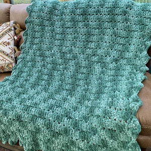 May include: A teal crocheted throw blanket with a scalloped edge. The blanket is made with a soft, plush yarn and has a subtle ombre effect.