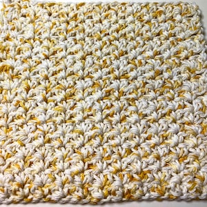 May include: A crocheted dishcloth in a white and yellow color scheme. The dishcloth is made with a textured stitch pattern.