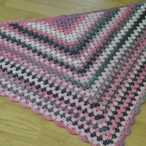 May include: A handmade crochet shawl in a triangular shape. The shawl features a gradient of pink, gray, and white colors. The pattern is a classic granny stitch, with a scalloped edge. The yarn has a subtle sparkle.