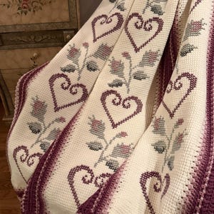 May include: A cream-coloured crocheted blanket featuring a repeating pattern of dark purple hearts and muted green floral stems. The blanket is finished with a thick, textured dark purple border along the edges, showcasing intricate stitch work.