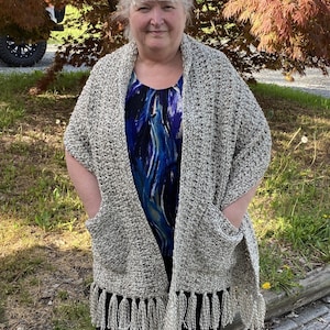 以下が含まれることがあります： フリンジが付いたベージュと白のクロシェ編みショール。ショールには両側にポケットが2つ付いています。