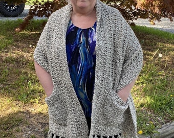 May include: A beige and white crocheted shawl with fringe. The shawl has two pockets on the sides.