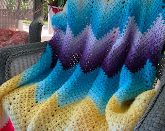 May include: A colorful crocheted blanket with a chevron pattern in shades of pink, yellow, blue, and purple. The blanket is draped over a wicker chair on a porch.