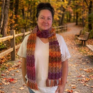 May include: A crochet scarf featuring a textured bobble stitch pattern in a variegated color palette of deep purple, burnt orange, brown, and gold. The scarf has a long rectangular shape with fringe detailing at both ends, worn over a plain white t-shirt.