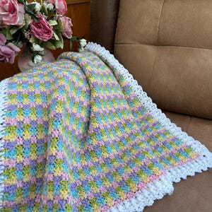 May include: A handmade, colorful crochet blanket with a white scalloped edge. The blanket features a pattern of pastel squares in shades of yellow, blue, green, and purple. The blanket is draped over a brown couch.