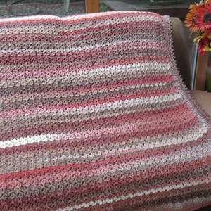 Peut inclure: Une couverture en crochet à rayures dans les tons de brun, rose et blanc. La couverture est posée sur un canapé en cuir marron.