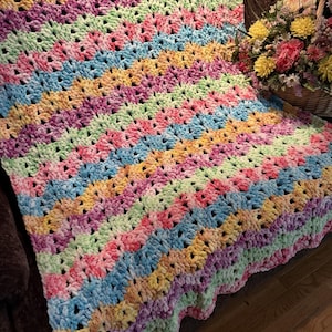 May include: A handmade crochet blanket featuring a wavy, chevron-style pattern. The yarn is a soft, plush texture in a repeating color palette of pastel pink, light blue, pale yellow, and mint green, creating a vibrant, multi-colored striped design.