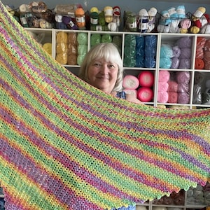 May include: A colorful, hand-knitted shawl with a striped pattern in shades of green, yellow, pink, and purple. The shawl is held up, showcasing its size and design. The background features shelves filled with various colors of yarn.