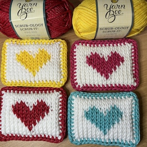 May include: Four handmade, rectangular crochet scrubbies with heart designs in yellow, pink, red, and teal. Each scrubby has a white center and a colored border. Two skeins of Yarn Bee Scrub-ology yarn in Fire Red and Lemon are in the background.