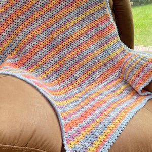 May include: A handmade crochet blanket in pastel colors. The blanket features a striped pattern with shades of orange, yellow, pink, and blue. The edges are finished with a light blue border. The blanket is draped over a brown armchair.