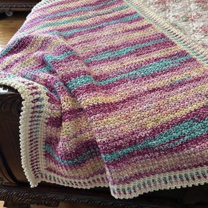 May include: A handmade crochet blanket with horizontal stripes in shades of purple, teal, yellow, and cream. The blanket has a cream-colored border and is draped over a bed. The blanket is a cozy home decor item.