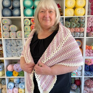 Puede incluir: Una mujer lleva una chal de ganchillo rosa y blanco con un borde blanco de ganchillo. El chal está drapeado sobre sus hombros y ella está sosteniendo los bordes del chal en sus manos. La mujer está de pie frente a una pared de hilo.