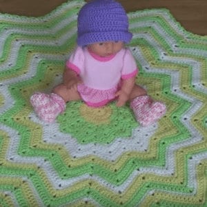 May include: A green, white, and yellow crocheted baby blanket with a starburst pattern. A baby doll is sitting on the blanket wearing a purple hat, pink shirt, and pink booties.
