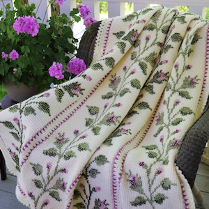 May include: A cream-colored, floral-patterned blanket draped over a wicker chair. The blanket features a repeating design of green leaves and purple flowers. A pot of pink flowers sits nearby, adding a touch of color to the scene.