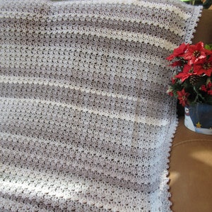 Peut inclure: Une couverture en crochet avec des rayures alternées de blanc et de gris. La couverture est pliée et posée sur un canapé brun.