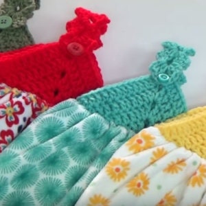 May include: Close-up of several colorful crochet and fabric kitchen towel toppers. The toppers are in various colors, including red, teal, and yellow, with button closures. The fabric towels have floral and geometric patterns.