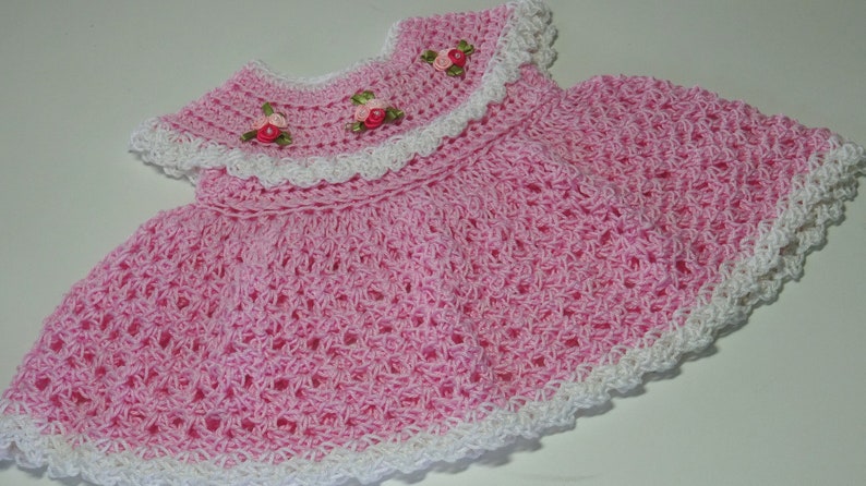 May include: A pink crocheted baby dress with white lace trim and pink ribbon flowers.