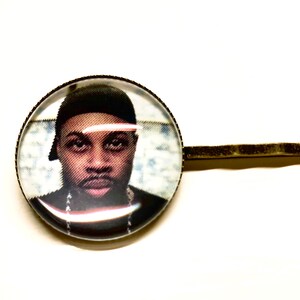 May include: A round glass hair clip with a photo of a man wearing a black hat and a gold chain. The clip is attached to a gold metal bar.
