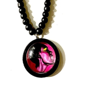 Könnte beinhalten: Eine schwarze Perlenkette mit einem runden Anhänger, der ein pinkfarbenes und gelbes Porträt zeigt.