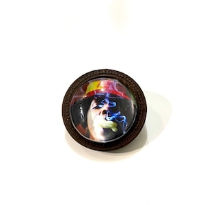May include: A round wooden pin with a glass insert featuring a colourful image of a person smoking a cigarette. The person is wearing a red and yellow hat.