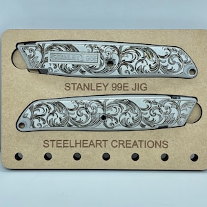 Op de afbeelding: Twee zilveren Stanley 99E utility messen met een gegraveerd bloemenpatroon. De messen worden getoond in een houten jig met de tekst "STEELHEART CREATIONS" eronder gedrukt.