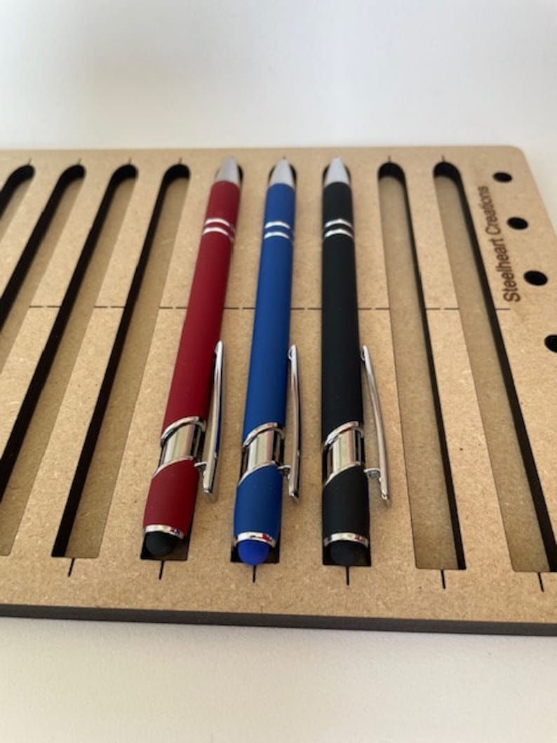 May include: Three pens in a wooden holder. The pens are red, blue, and black. The holder has the text "Steelheart Creations" on it.