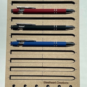 May include: A wooden jig with slots for holding ink pens. The jig has the text "Ink Pen Jig" and "Steelheart Creations" printed on it.