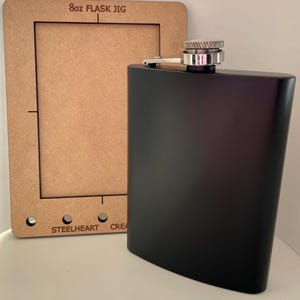 Puede incluir: Una petaca negra con tapón y bisagra plateados se encuentra junto a una plantilla de madera. La plantilla tiene el texto "8oz FLASK JIG" y la marca "STEELHEART CREA". La petaca tiene forma curva y mide unos 14 cm de alto.