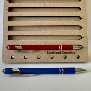 May include: A wooden pen holder with slots for pens. The holder has a red pen and a blue pen in it. The holder has the text "Steelheart Creations" printed on it.