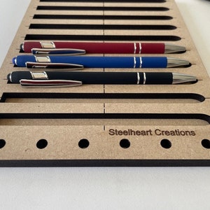 May include: A wooden pen holder with slots for storing pens. The holder has a black border and the text "Steelheart Creations" is printed on the front.