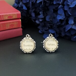 Peut inclure: Deux pendentifs argentés avec un bord floral, chacun avec du texte d'une page de livre. L'un dit "I think Atreyu, while" et l'autre "wered Artax, like a w". Des livres rouges et des fleurs bleues sont en arrière-plan.