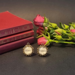 May include: Two round, antique-style pendants with text from a book, featuring the words "Mary" and "Poppins". The pendants are set in a bronze-colored metal with a floral edge. A stack of red books and red roses are in the background.