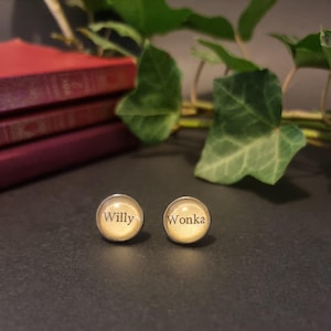 May include: A pair of round stud earrings with the words "Willy Wonka" printed on them. The earrings have a silver-colored border and a cream-colored background. The background includes a stack of red books and green leaves.