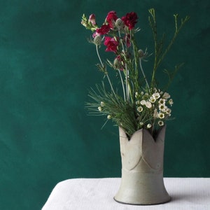 Peut inclure: Un vase en céramique blanche avec un bord en forme de couronne, rempli d'un bouquet de fleurs roses et blanches. Le vase est posé sur une nappe blanche sur un fond vert foncé.