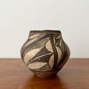 May include: A small, rounded ceramic pot featuring a cream-colored base with bold, black geometric patterns. The design consists of sharp, angular shapes and fine, parallel line details. The base of the vessel shows a natural, unpainted clay texture.
