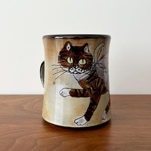 May include: A ceramic mug with a whimsical cat design. The cat is brown with black stripes and white paws, set against a beige background. The mug has a dark brown rim and handle, and is approximately 4 inches tall.