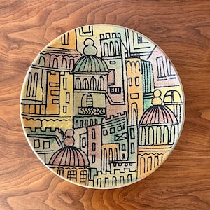 May include: A round decorative plate featuring a colorful cityscape design. The plate showcases buildings with domes and arches in shades of yellow, orange, green, and blue, outlined in black. The plate is set against a wooden surface.
