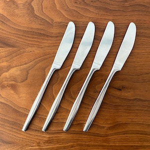 May include: Four stainless steel butter knives with rounded blades and long, slender handles are arranged diagonally on a wooden surface. The knives have a polished, reflective finish, and the wood grain is visible.