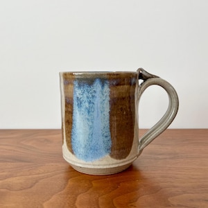 May include: A white ceramic mug with a black handle and the text "But First, Coffee" in a bold, black font. The mug is set against a blurred background, suggesting a cozy setting.