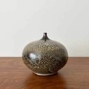 May include: A speckled ceramic vase with a rounded body and a narrow neck. The vase has a dark brown and tan speckled pattern, with a glossy finish. The vase is sitting on a wooden surface.