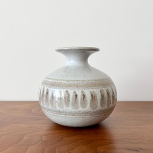 May include: A small, round, off-white ceramic vase with a narrow neck and a decorative band of vertical, teardrop-shaped brown patterns around the middle. The vase sits on a wooden surface.