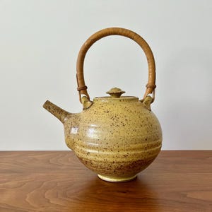 May include: A ceramic teapot with a speckled, yellow-brown glaze. It features a rounded body, a spout, and a woven handle. The teapot is displayed on a wooden surface against a neutral background, showcasing its rustic design.