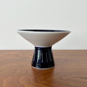 May include: A modern, ceramic bowl with a white interior and a navy blue rim. The bowl has a flared, conical shape, supported by a navy blue, cylindrical base with vertical grooves. The bowl sits on a wooden surface.