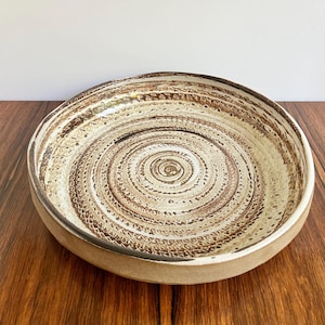 May include: A ceramic bowl with a spiral design in shades of brown and cream. The bowl has a wide, shallow shape and a textured surface. The rim is a solid cream color, and the bowl sits on a wooden surface.