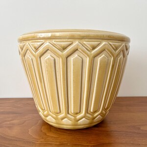 May include: A glossy, mustard-yellow ceramic planter featuring a repeating geometric pattern of vertical panels and hexagonal shapes. The vessel has a wide, circular opening and tapers toward a rounded base, with a smooth, reflective finish.