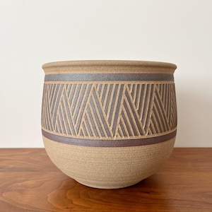 May include: A beige ceramic planter with a geometric pattern. The planter has a rounded shape with a textured surface. The design features a series of angled lines in a darker brown color, creating a visual contrast. A dark brown band encircles the base.
