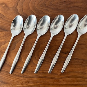 May include: Six stainless steel spoons with elongated oval bowls and slender handles are arranged on a wooden surface. The spoons are reflective, and the wood grain provides a warm contrast. A set of elegant flatware.
