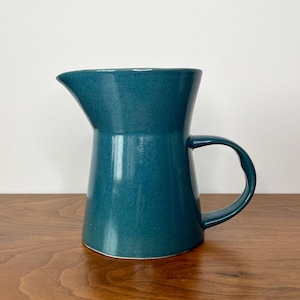 May include: A teal ceramic pitcher with a curved handle and a flared spout. The pitcher has a glossy finish and sits on a wooden surface. The design is simple and elegant, suitable for serving beverages.