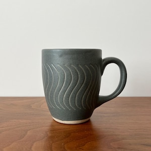 May include: A dark gray ceramic mug with a curved handle. The mug features a wave pattern carved into the surface. The mug sits on a wooden surface. The mug is approximately 4 inches tall.